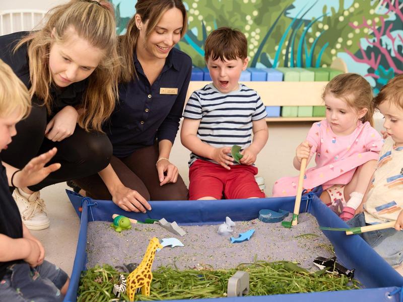 Kinder spielen mit Spielzeugtieren in einem Sandkasten, zwei Erwachsene beobachten freundlich.