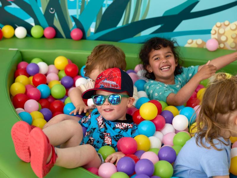 Drei Kinder spielen lachend in einem bunten Bällebad mit farbigen Plastikkugeln.