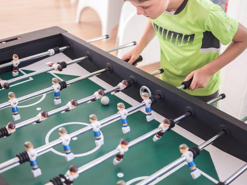 Kind spielt Tischfußball an grünem Spielfeld mit kleinen Kunststoffspielern in blauer und roter Kleidung.