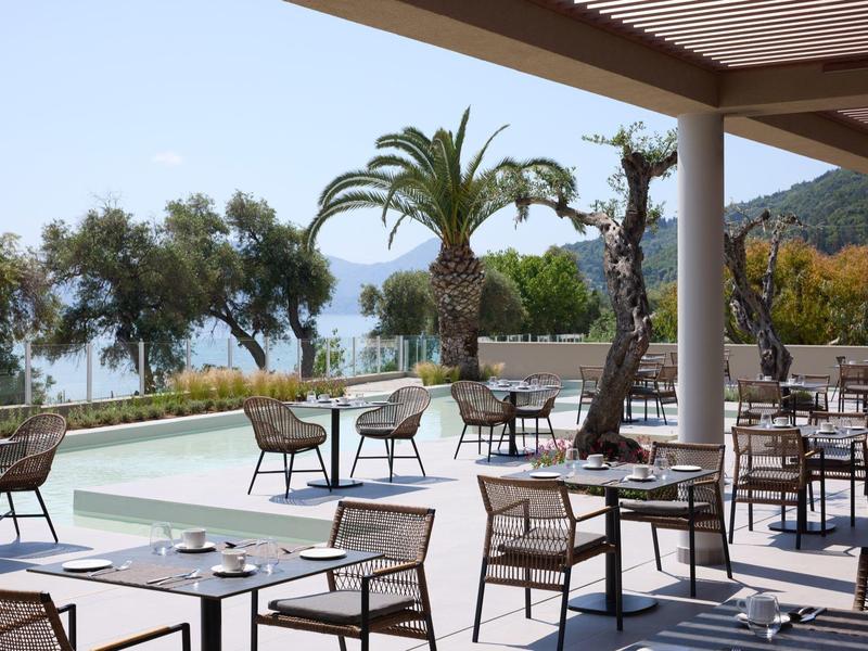 Moderner Außenbereich mit Tischen, Stühlen und Palmen, Blicke auf Meer und Berge im Hintergrund.