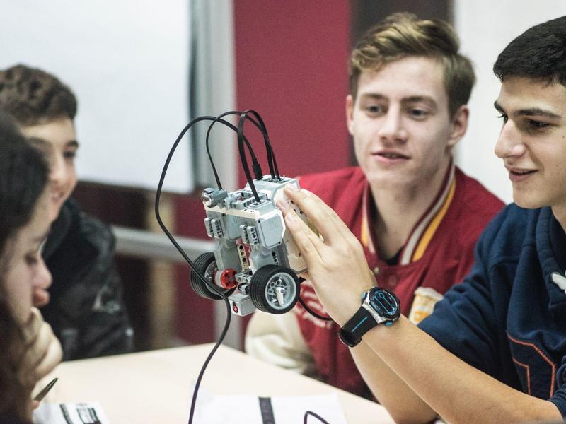 Vier Jugendliche betrachten gespannt einen kleinen grauen Roboter mit Rädern an einem Tisch.