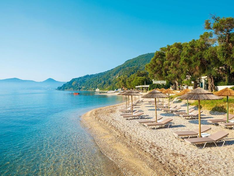 Strand mit Kies, Sonnenschirmen und Liegen, dahinter grüne Hügel und blauer Himmel.