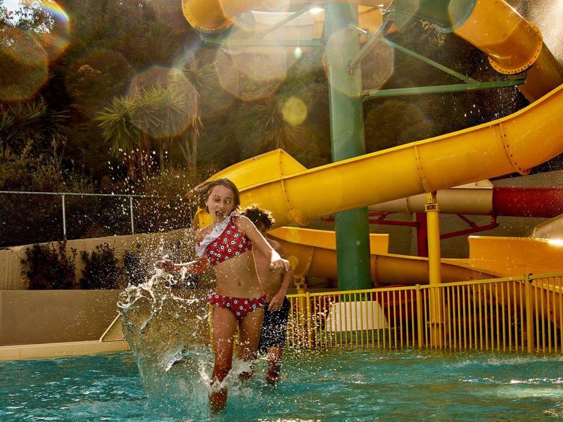 Kind springt ins Wasser, im Hintergrund gelbe Wasserrutsche, Sonne strahlt durch Bäume.