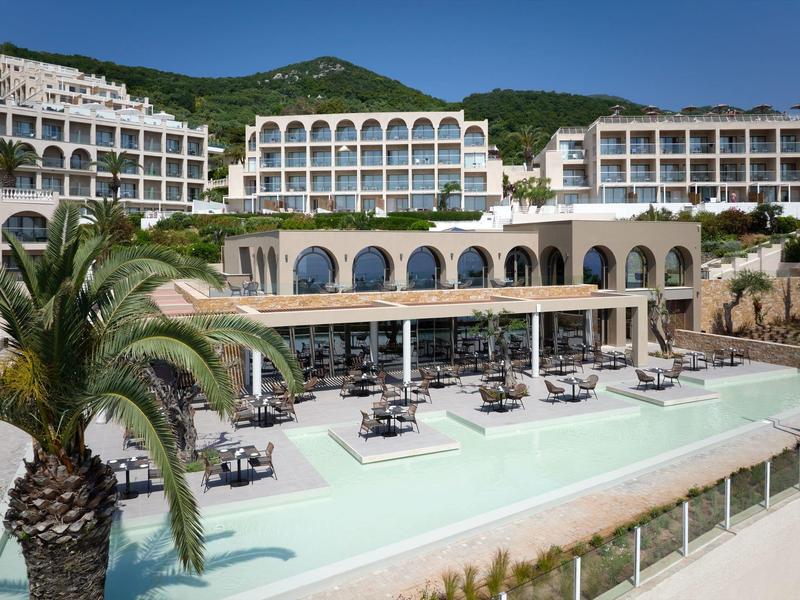 Großes Hotel mit mehreren weißen Gebäuden, Pool mit Sonnenschirmen und Palmen davor, grüner Berg im Hintergrund.