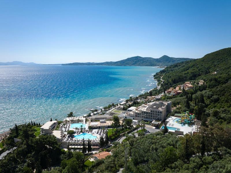 Blick auf eine Küstenanlage mit Pools, grünen Hügeln und klarem blauem Meer unter blauem Himmel.