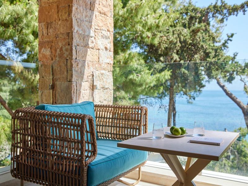 Gemütliche Terrasse mit blauem Korbsessel und Holztisch vor See, umgeben von Bäumen und Steinmauer.