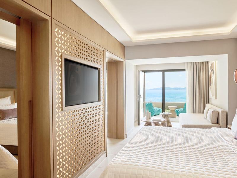 Helles Hotelzimmer mit Doppelbett, Sofa, Meerblick und dekorativer Holzwand mit Fernseher.