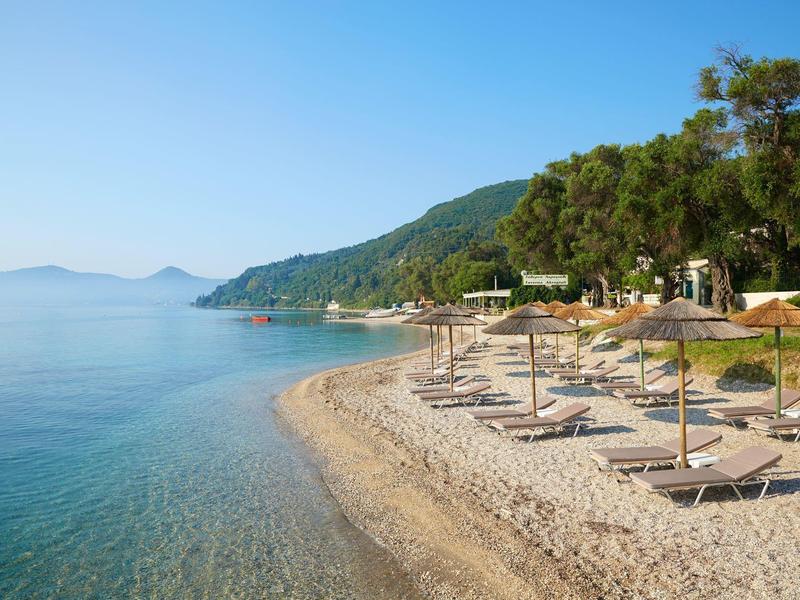 Strand mit Kieselsteinen, Liegen und Sonnenschirmen, umgeben von grün bewaldeten Hügeln.
