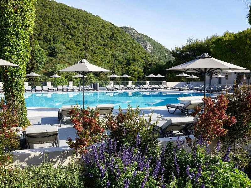 Großer Pool mit Liegestühlen, Sonnenschirmen und grünen Bergen im Hintergrund bei klarem Himmel.