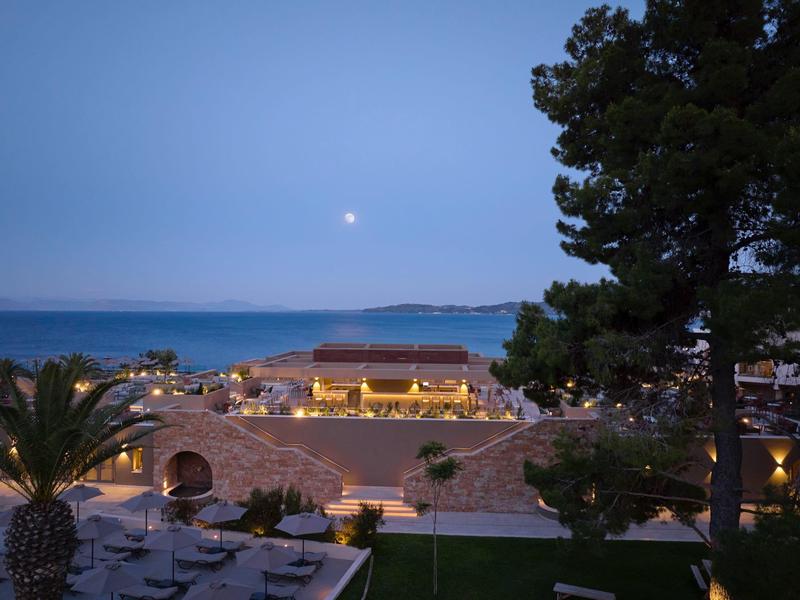 Abendlicht über einer Küstenstraße mit beleuchteten Gebäuden, Bäumen und ruhigem Meer im Hintergrund.