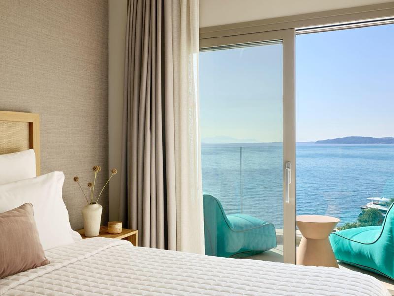 Gemütliches Zimmer mit Bett, Kissen, Pflanzen und Balkon mit Blick auf das blaue Meer.