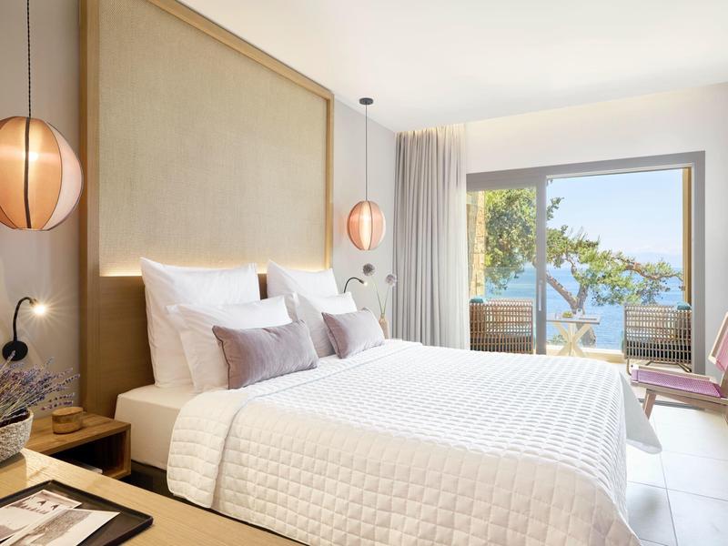 Helles Schlafzimmer mit großem Bett, weißen Bettdecken, zwei runden Lampen und Blick auf Terrasse und Meer.