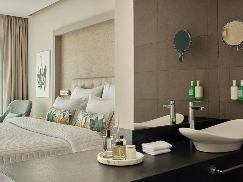 Modernes Schlafzimmer mit großem Bett, heller Farbgestaltung und angrenzendem Bad mit Waschbecken.