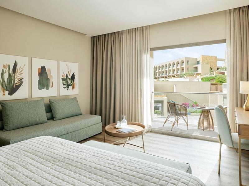 Helles modernes Schlafzimmer mit großem Bett, Grünsofa, Holztisch und Balkon mit Stühlen.