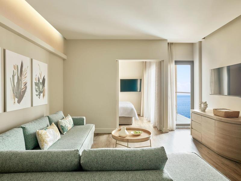Helles Wohnzimmer mit grünem Sofa, runden Tischen, Holzmöbeln und Blick auf Meer und Schlafzimmer.