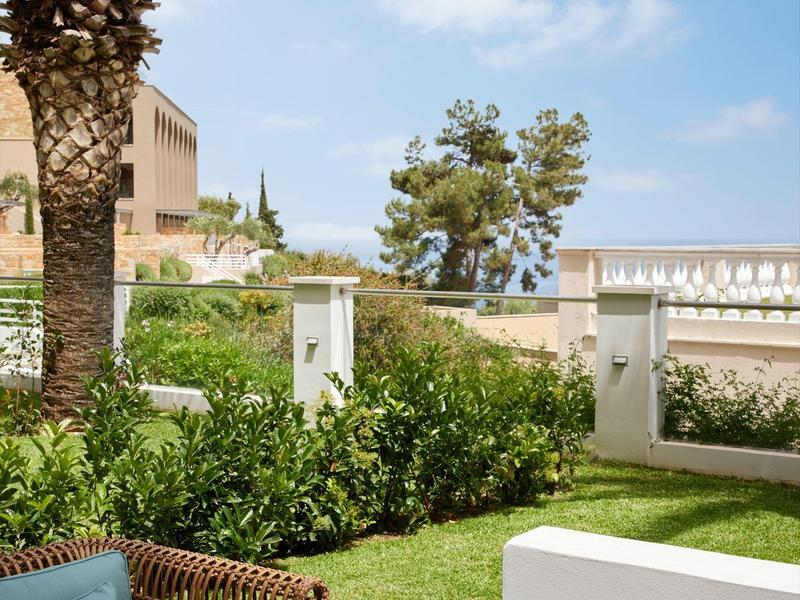 Garten mit grünen Pflanzen, Palmen, blauem Himmel, Holztisch und blauem Kissen auf Stuhl.