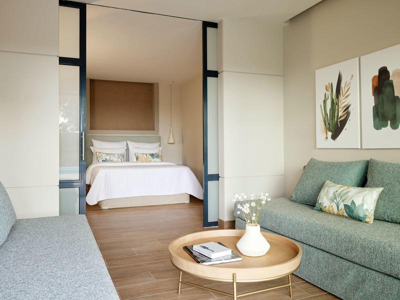 Helles Wohnzimmer mit grünem Sofa, rundem Holztisch und Blick auf ein Schlafzimmer mit weißem Bett.