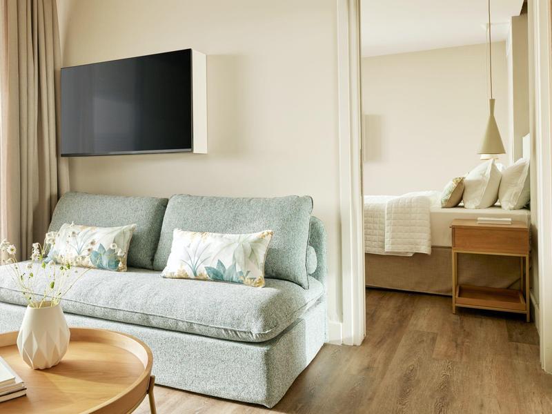 Helles Wohnzimmer mit blauem Sofa, Blumenmustern, Holzboden und Blick ins Schlafzimmer mit Bett.