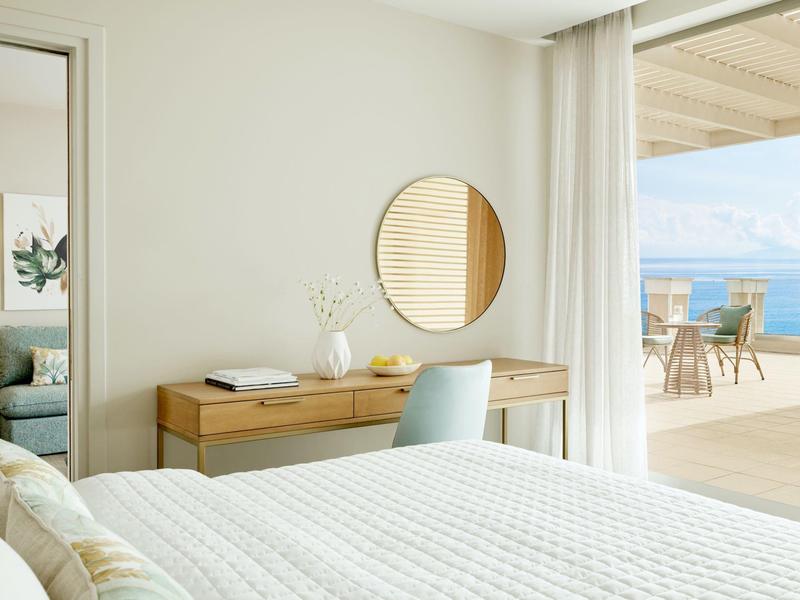 Helles Schlafzimmer mit weißer Bettdecke, Holzschreibtisch, rundem Spiegel und Zugang zur Terrasse.