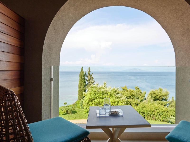 Blick durch großen Bogen auf Garten mit See, moderner Wohnraum mit Holzwand und hellem Tisch.