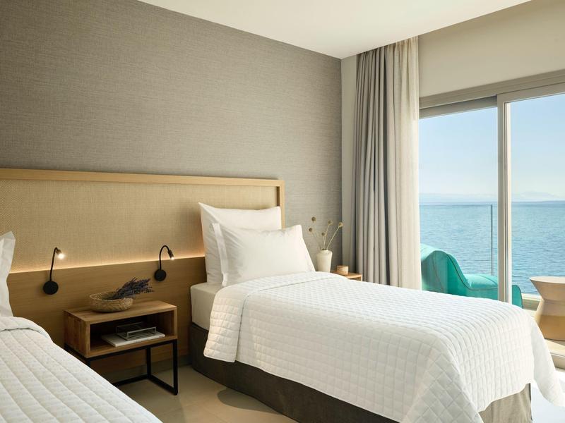 Helles Hotelzimmer mit zwei Einzelbetten, Nachttisch, Leselampen und Meerblick durch großes Fenster.