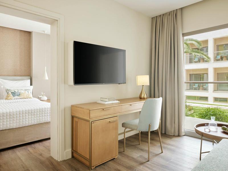 Moderner Raum mit Schreibtisch, Stuhl, großem Fernseher, Bett im Nebenraum und großem Fenster.