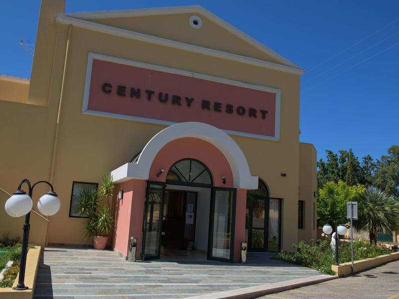 Gebäude mit gelber Fassade und rotem Bogen, Schild "Century Resort", Lampen und grüner Umgebung.