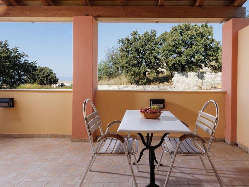 Terrasse mit Tisch, vier Metallstühlen und einem Obstkorb, umgeben von beigen Wänden und grünen Bäumen.