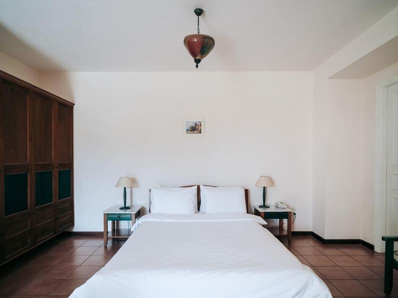 Schlichtes Schlafzimmer mit weißem Bett, Holzmöbeln, Nachttischen und braunem Boden.