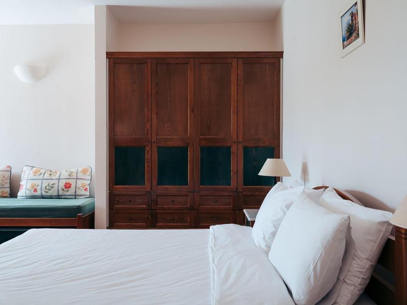 Schlafzimmer mit weißem Bett, Holzkasten, zwei Lampen, Sofa links und einem kleinen Bild an der Wand.