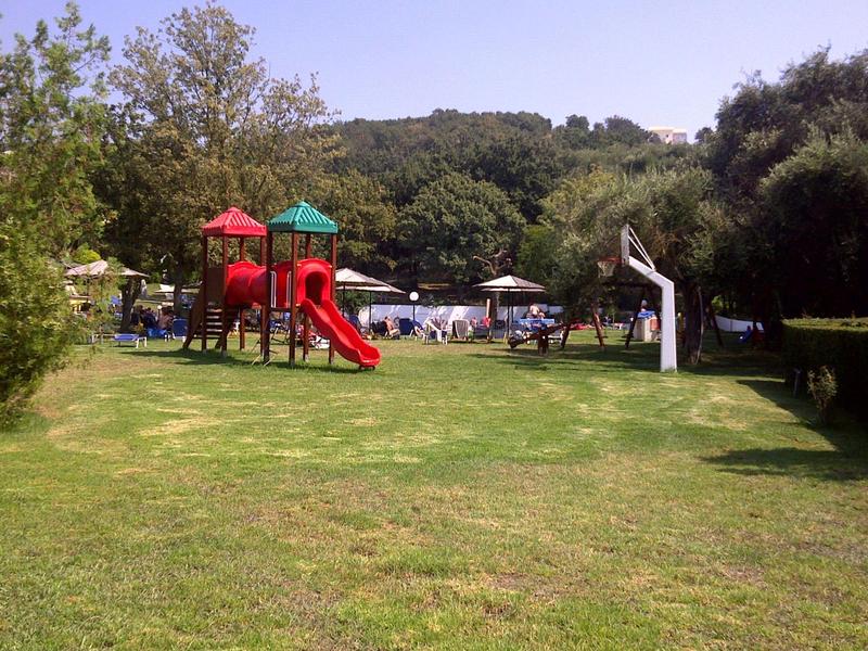 Spielplatz mit roten Rutschen und Klettergerüst auf grünem Rasen, umgeben von Bäumen und Hügeln.