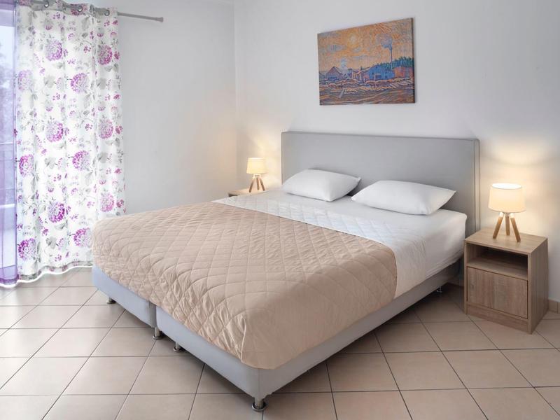 Modernes Schlafzimmer mit großem Bett, beigem Tagesdecke, Nachttisch und zarten lila Vorhängen.