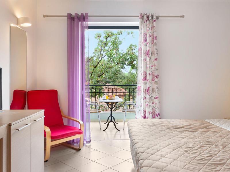 Helles Schlafzimmer mit beigem Bett, rotem Sessel und Balkon mit grünem Baum im Hintergrund.