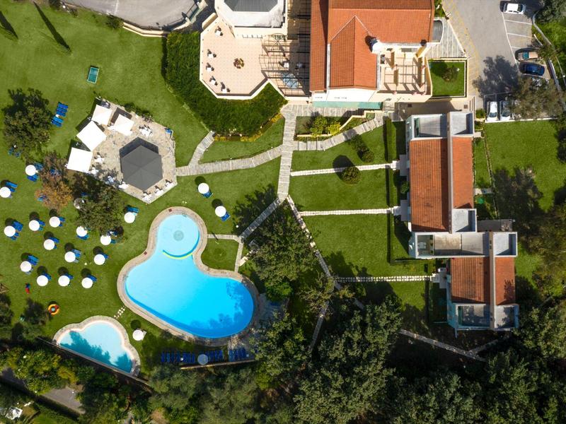 Luftaufnahme von Hotel mit blauem Pool, Sonnenschirmen, Wegen und grünen Wiesen in Brunchform.