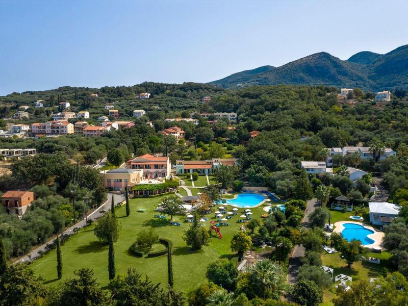 Grüner Hügel mit mehreren Gebäuden, blauen Pools und Bergen im Hintergrund unter blauem Himmel.