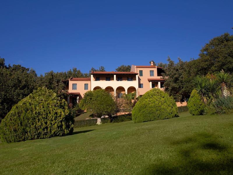 Großes rosa Haus mit Arkaden auf Hügel, umgeben von grünen Büschen und Rasen unter blauem Himmel.