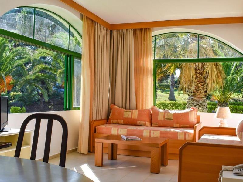 Helles Wohnzimmer mit großen grünen Fensterrahmen, orangefarbener Couch und Blick auf Palmen.