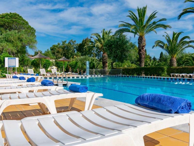 Leere weiße Liegestühle mit blauen Handtüchern am Pool, umgeben von Palmen unter blauem Himmel.