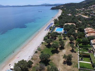 Langer Sandstrand mit klarem blauem Wasser, angrenzend an grüne Bäume und Ferienhäuser.