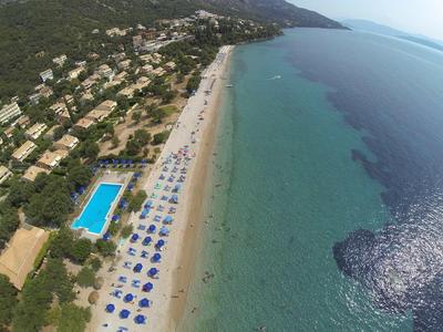 Luftaufnahme eines langen Sandstrands mit blauen Sonnenschirmen, einem Pool und Häusern in grüner Landschaft.