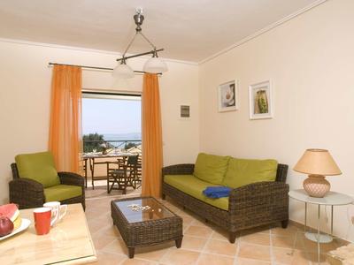 Helles Wohnzimmer mit Rattansofa, grün-gelben Kissen, Sessel, Glastisch und orangenen Vorhängen.