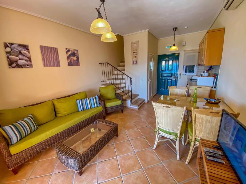 Gemütliches Wohnzimmer mit gelbem Sofa, Kamin, Treppe und offener Küche in warmen Farben.