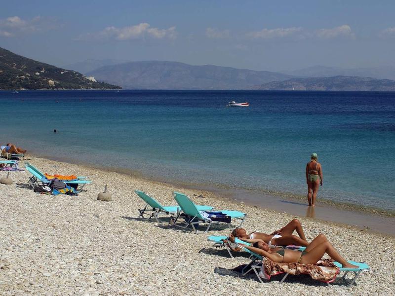 Kieselstrand mit Sonnenliegen, einigen Personen und ruhigem, blauem Meer unter hellem Himmel.