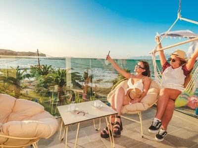 Dos mujeres se relajan en sillas de balcón con vista al mar en clima soleado.