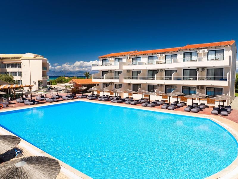 Großer Außenpool mit Sonnenliegen und Hotelgebäude unter klarem blauem Himmel.