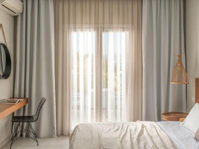 Schlichtes Schlafzimmer mit Doppelbett, hellen Vorhängen, Holzregal, schwarzem Stuhl und runder Spiegel.