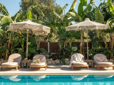 Vier Sonnenliegen mit Kissen und zwei weißen Sonnenschirmen am blauen Pool, umgeben von grünen Pflanzen.