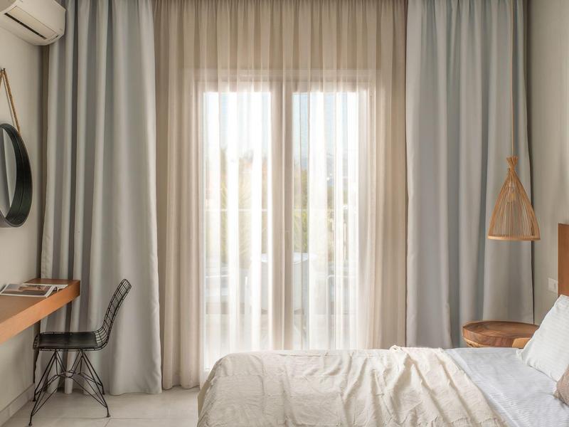 Schlichtes Schlafzimmer mit Doppelbett, hellen Vorhängen, Holzregal, schwarzem Stuhl und runder Spiegel.