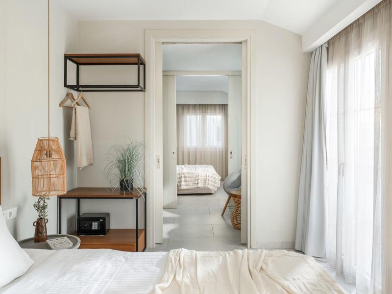 Helles Schlafzimmer mit weißer Bettwäsche, Holzmöbeln und großem Spiegel, daneben Gardinen.