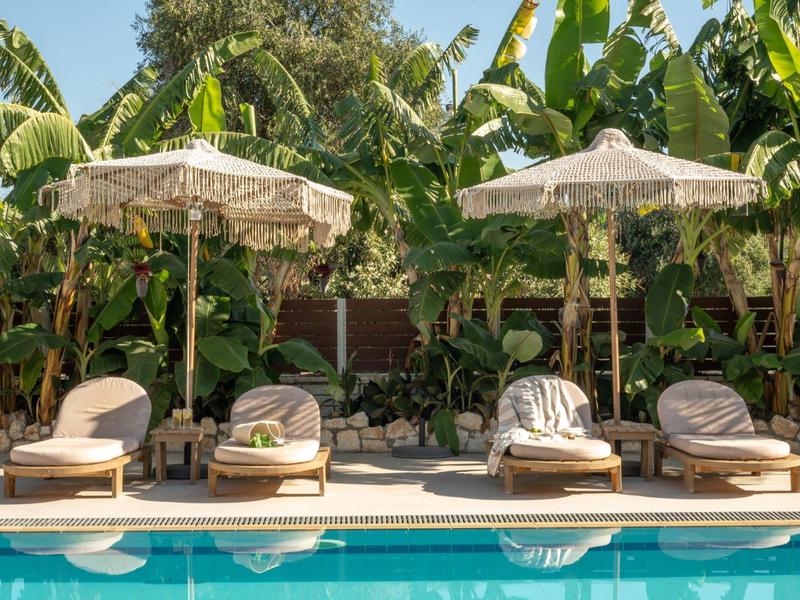 Vier Sonnenliegen mit Kissen und zwei weißen Sonnenschirmen am blauen Pool, umgeben von grünen Pflanzen.